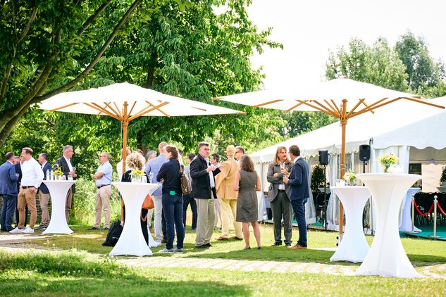 Verschiedene Personen stehen auf Rasen an Stehtischen unter Sonnenschirmen