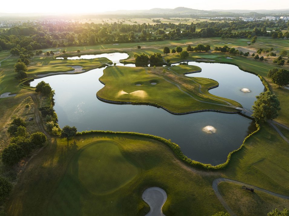 Platz St. Leon Luftaufnahme Inselgrün von Wasser umgeben bei Sonnenuntergang