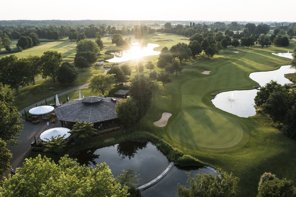 Luftaufnahme von flachem Gebäude mit Sonnenschirmen auf Terrasse neben Golfbahn und Teich