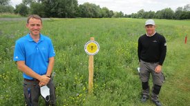 Daniel Lüttger und Karsten List stehen vor Blumenwiese mit gelbem Lebensraum Golfplatz Schild